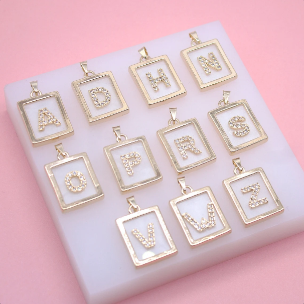 A collection of gold rectangular initial letter pendants encrusted with sparkling cubic zirconia crystals rests on a white display against a pink background.