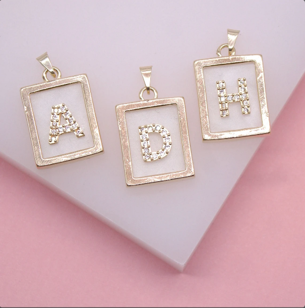 Three gold rectangle initial pendants featuring sparkling cubic zirconia letters A, D, and H rest on a white and pink surface.