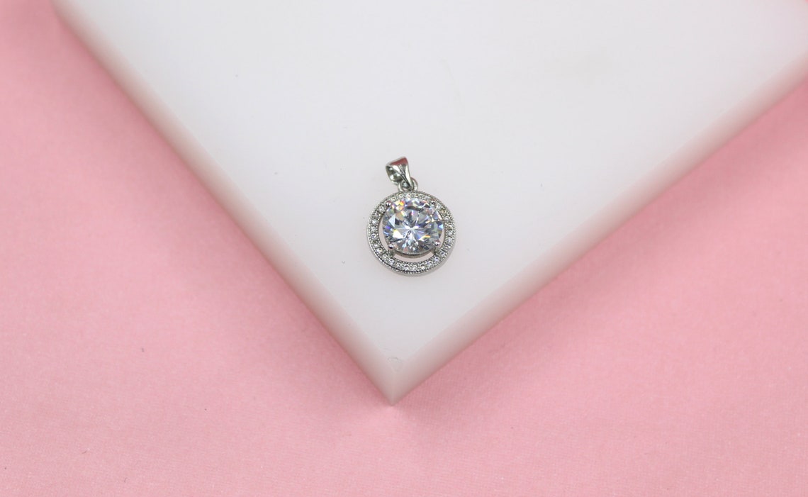 A sparkly silver halo diamond pendant rests on a white display against a soft pink background.