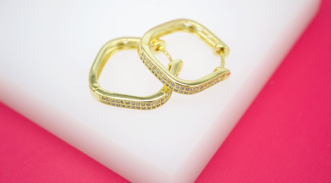 Two gold geometric hoop earrings embellished with pavé diamonds rest on a white display surface against a vibrant pink background.