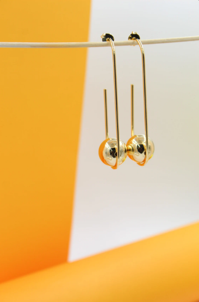 A pair of modern gold hoop earrings featuring geometric shapes and spherical charms hangs gracefully against a bright orange and white backdrop.