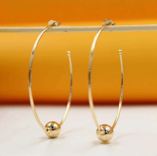 A pair of elegant gold hoop earrings with polished bead charms hangs suspended on a thin stick against an orange and white background.