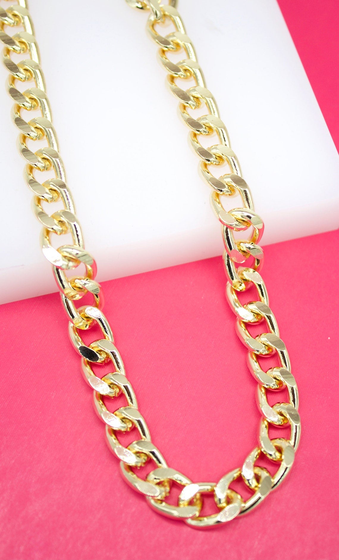 A chunky gold curb chain necklace rests against a vibrant pink and white background.