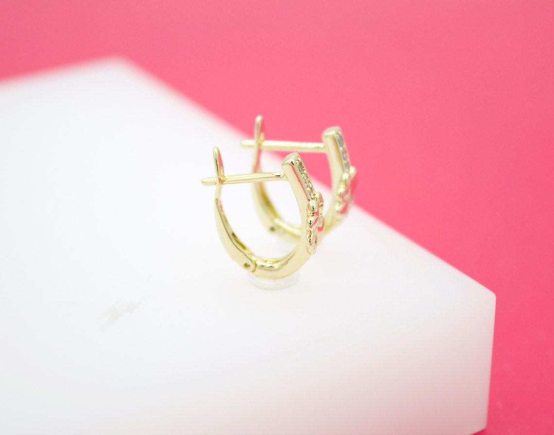 A pair of gold huggie hoop earrings with sparkling crystal accents rests on a white display surface against a vibrant pink background.
