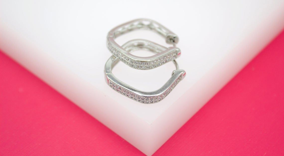 A pair of geometric pave hoop earrings rest on a white display block against a vibrant pink background.