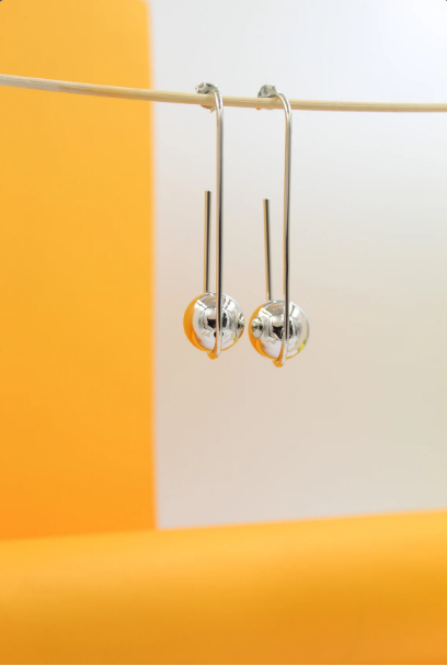 A pair of modern silver geometric drop earrings featuring hanging spheres dangles delicately against a vibrant yellow and white backdrop.