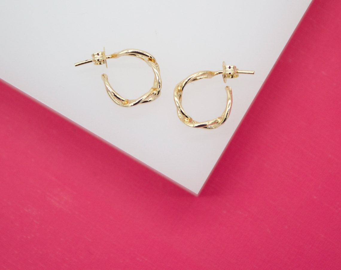 A pair of elegant twisted gold hoop earrings rest on a white surface against a bright pink background.