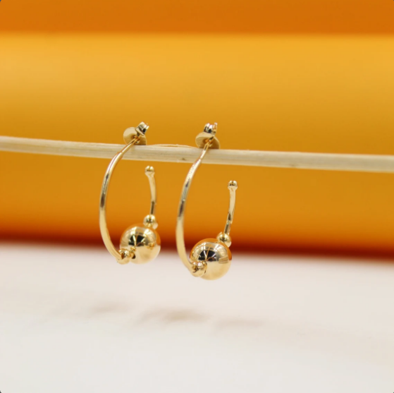 A pair of shiny gold hoop earrings featuring dangling bead charms hangs delicately from a wooden stick against an orange and white background.