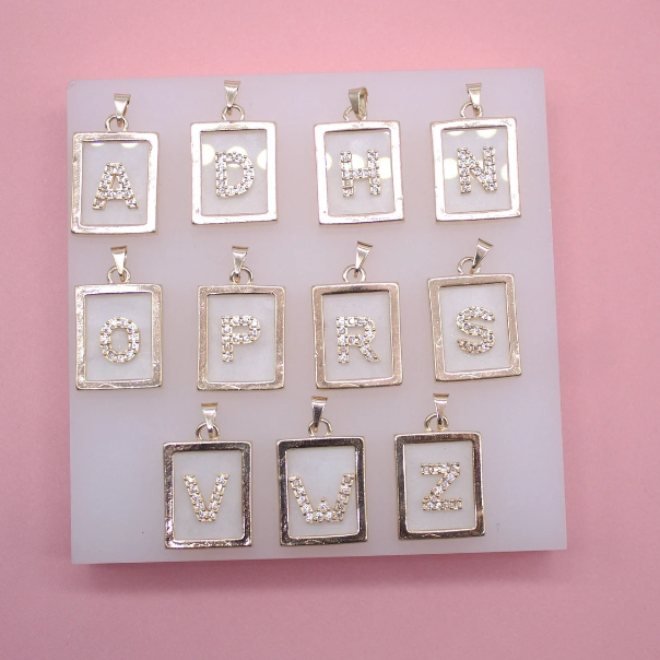A collection of gold-framed initial pendant necklaces with rhinestone-encrusted letters is displayed on a white board against a pink background.