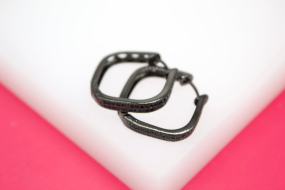 A pair of black geometric hoop earrings rests on a white display surface against a pink background.