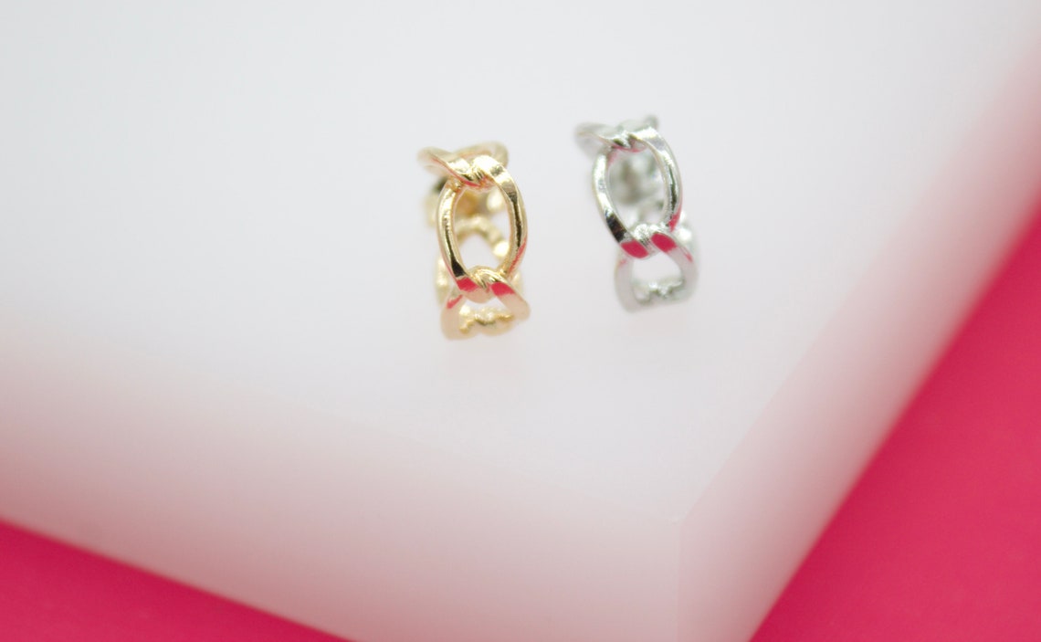 A gold hoop earring and a silver hoop earring featuring an intertwined chain design rest on a white surface against a pink background.