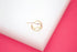 A delicate gold hoop earring rests on a white display surface against a vibrant pink background.