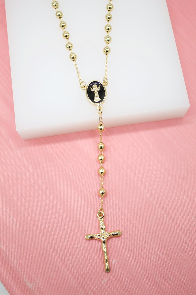 A gleaming gold rosary necklace with polished beads, a black centerpiece, and a crucifix rests on a white display block against a soft pink background.