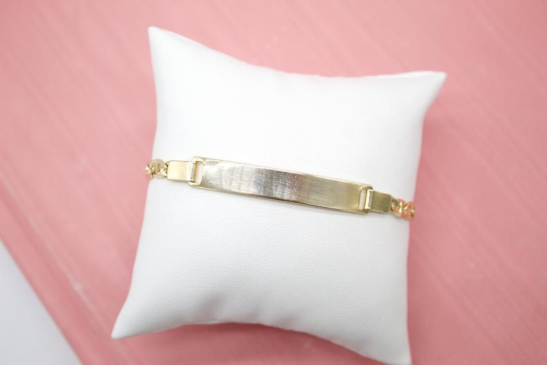 A gold ID bracelet rests on a small white display pillow against a pink background.