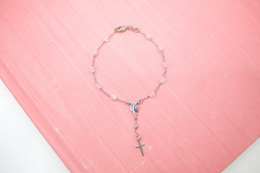 A delicate rosary bracelet featuring clear crystal beads and a silver crucifix rests on a soft pink surface.