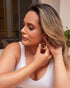 A woman with blonde-streaked hair wears a white top and puts on large, geometric gold dangle earrings.