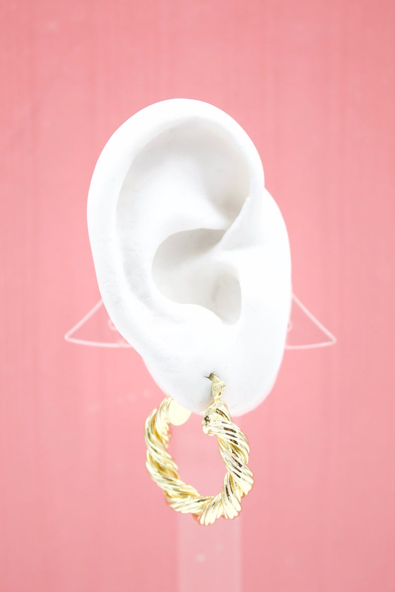 A white mannequin ear displays a shiny gold twisted hoop earring against a soft pink background.