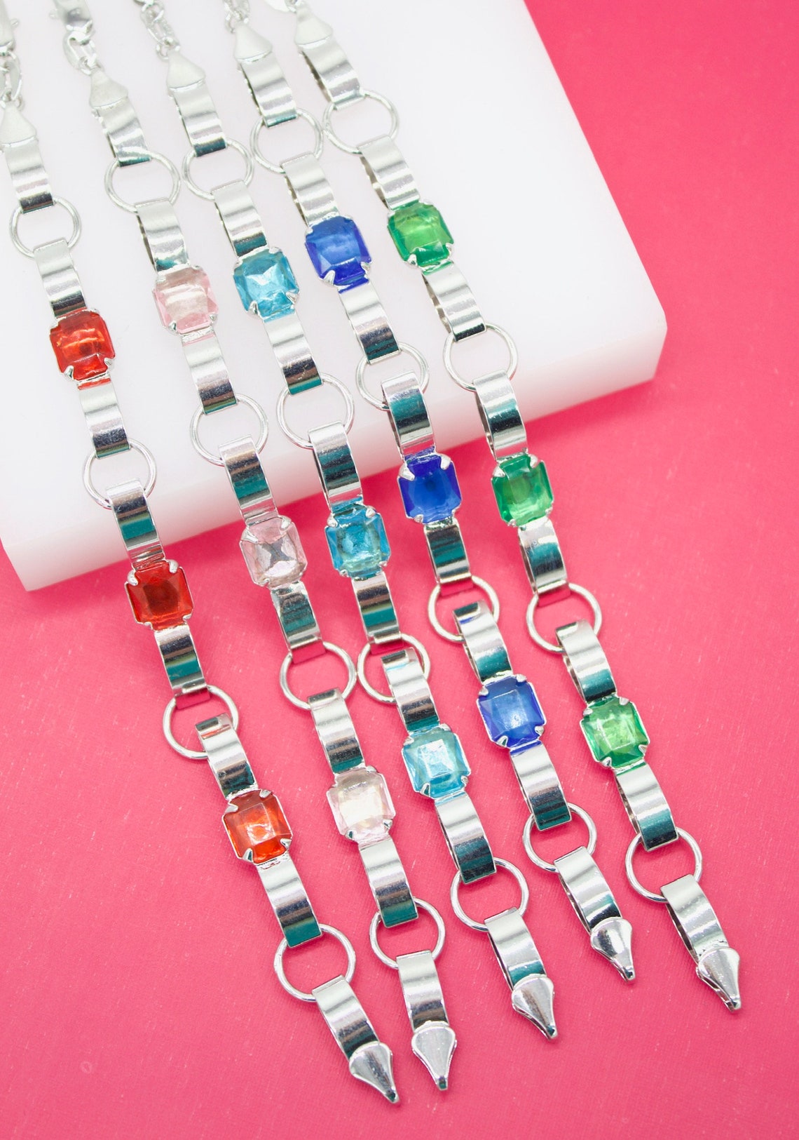 Five silver-tone clip-on suspenders, featuring colorful crystal rhinestone accents, rest neatly on a white display block against a vibrant pink background.