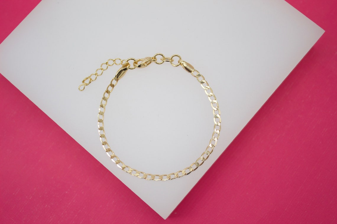 A delicate gold-plated curb chain bracelet rests on a white display surface against a bright pink background.