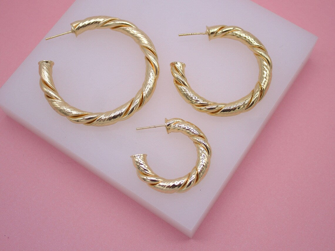 A set of three twisted gold hoop earrings rests on a white display block against a pink background.