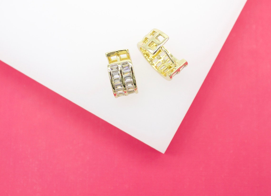 A pair of gold huggie hoop earrings embedded with baguette crystals rests on a white and pink surface.