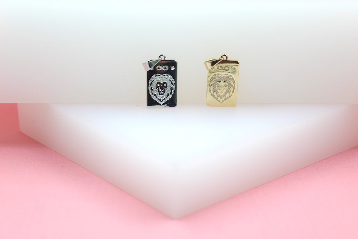 Two rectangular lion-engraved pendants, one silver and one gold, rest on a white display surface against a pink background.