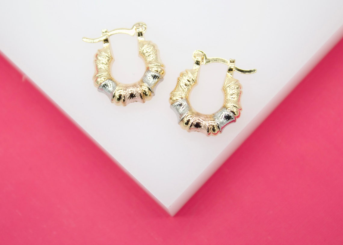A pair of textured tricolor gold bamboo hoop earrings rests on a white display surface against a vibrant pink background.