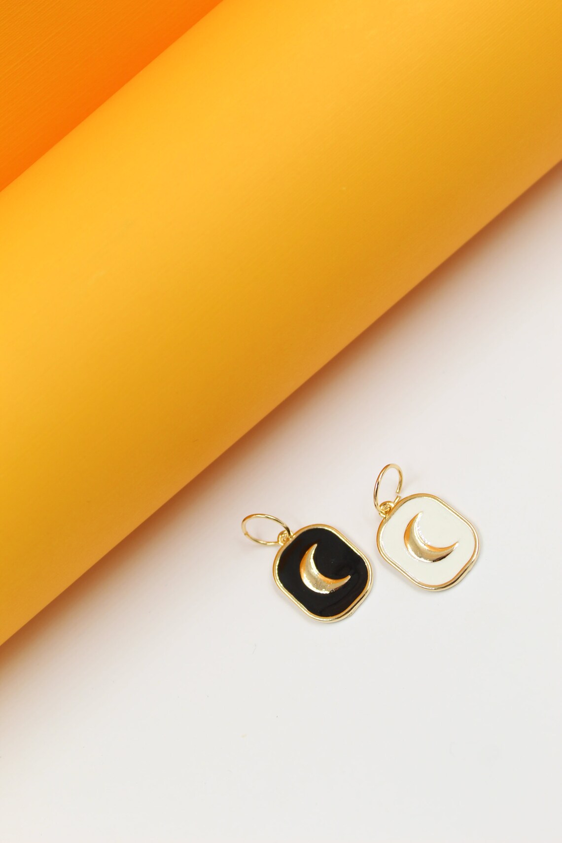 Two celestial charm pendants featuring a gold crescent moon on black and white enamel rest against a white surface beneath a vibrant yellow backdrop.