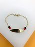 A gold ID bracelet rests on a white display stand, featuring delicate links, an engravable plaque, and accents of red and black beads.