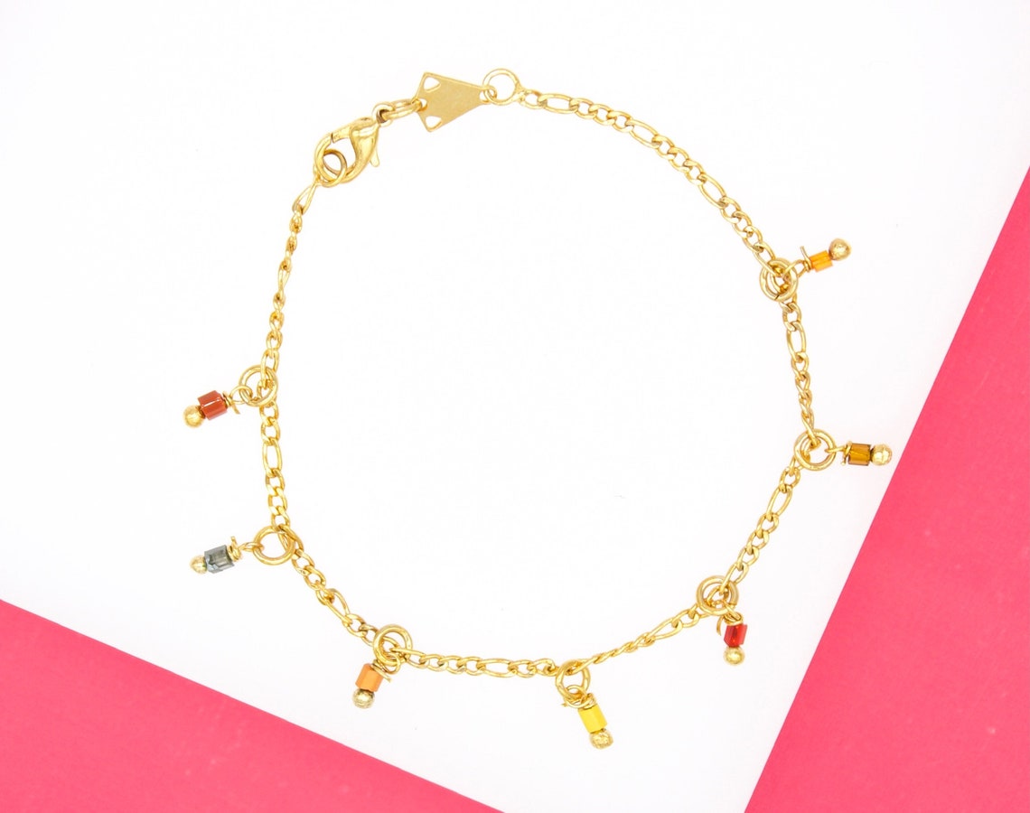 A delicate gold charm bracelet displays colorful, dangling bead accents against a pink and white background.