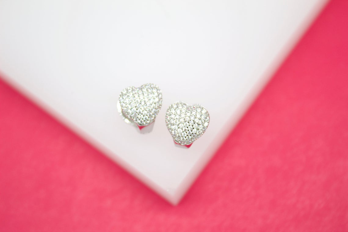 A pair of shimmering silver heart earrings encrusted with small pave diamonds rests on a white display surface against a vibrant pink background.
