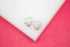 A pair of shimmering silver heart earrings encrusted with small pave diamonds rests on a white display surface against a vibrant pink background.