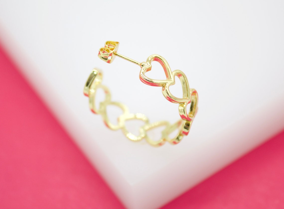 A gold hoop earring featuring an interconnected heart design rests against a pink and white background.