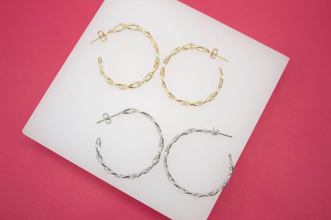 Two pairs of twisted metal hoop earrings in gold and silver rest neatly on a white display block against a vibrant pink background.