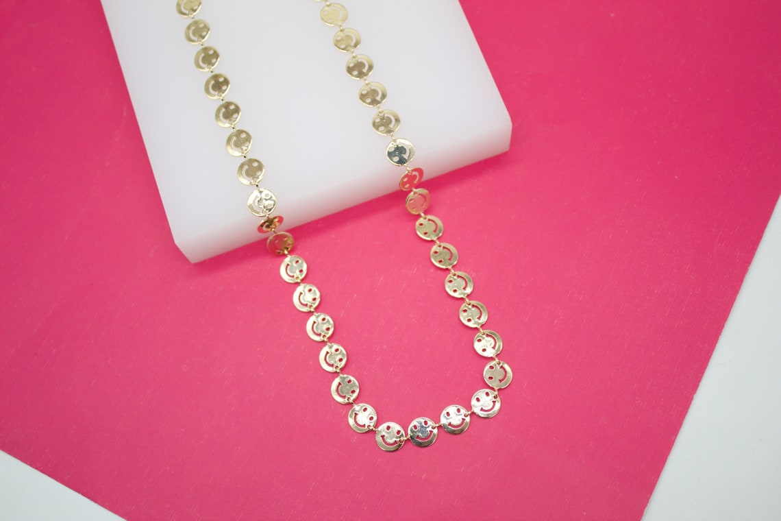 A gold smiley face necklace rests on a white display block against a vibrant pink background.