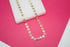 A gold smiley face necklace rests on a white display block against a vibrant pink background.