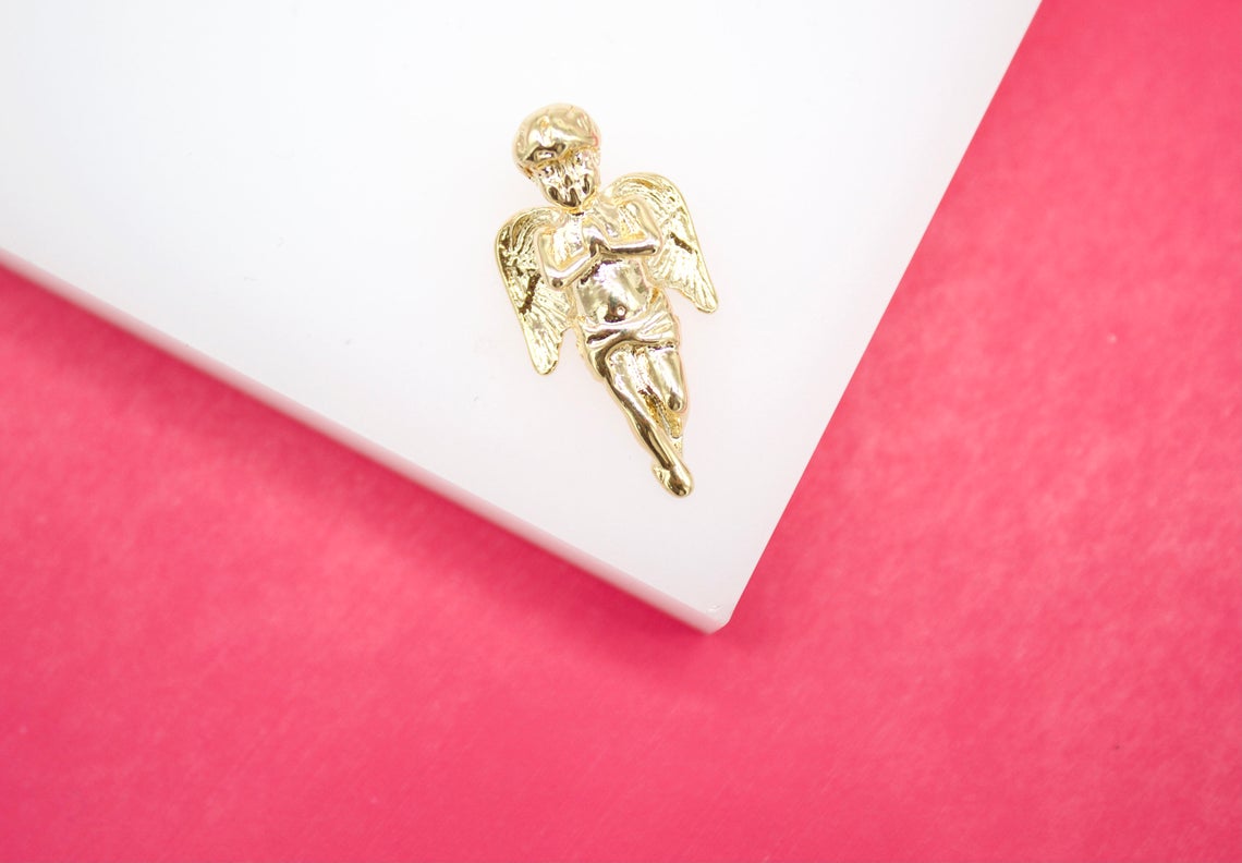 A shiny gold cherub pendant rests on a white display surface against a vibrant pink background.