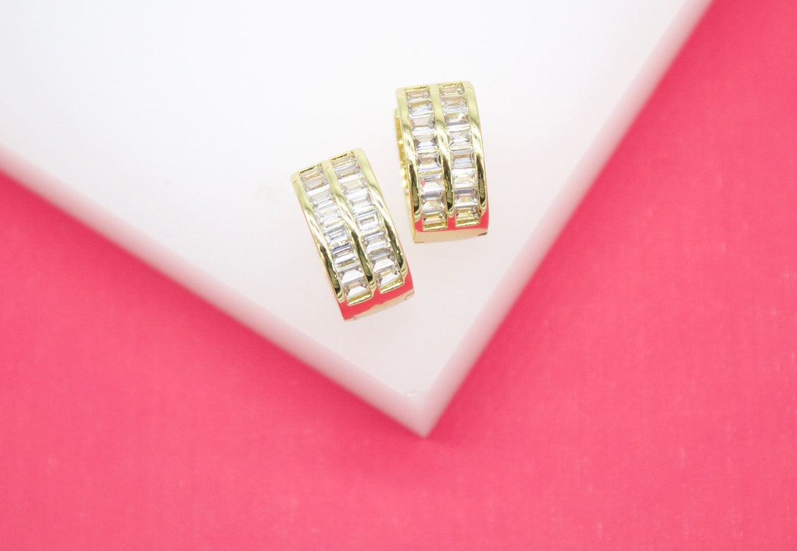 A pair of gold huggie hoop earrings featuring sparkling baguette crystals rests on a white and pink display surface.
