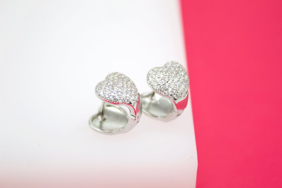 A pair of glittering pave-set heart earrings rests on a white and pink display stand.