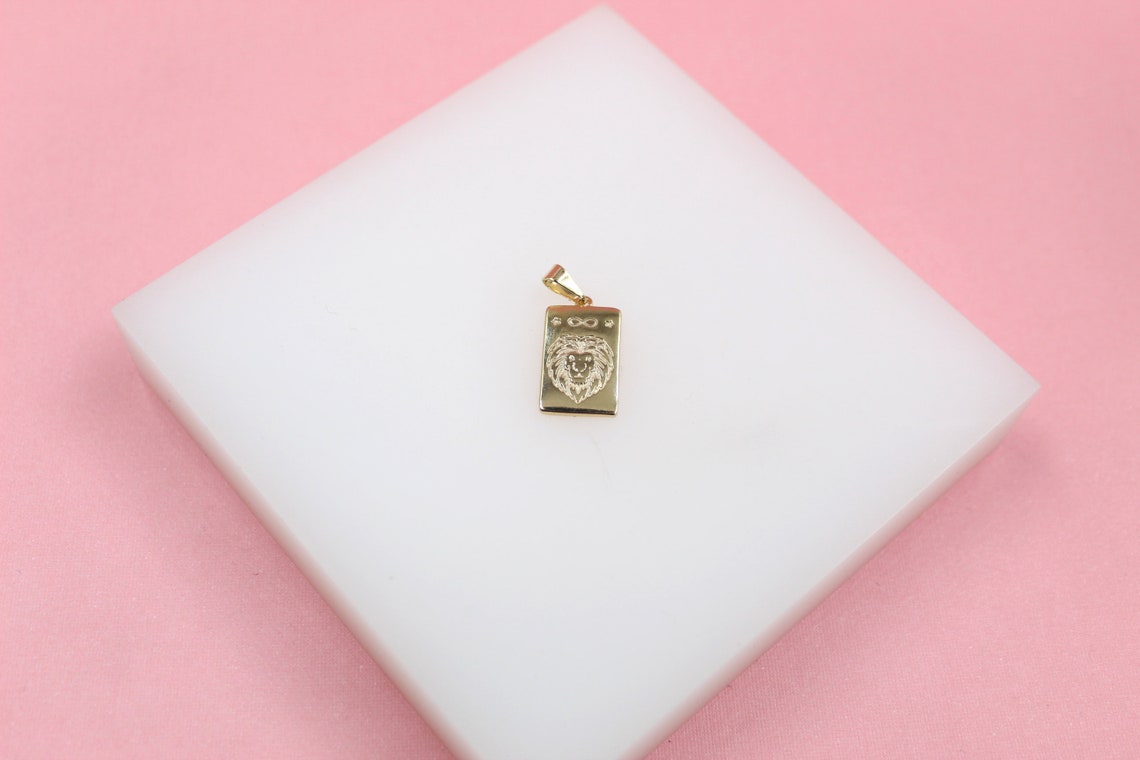 A shiny gold rectangular pendant featuring an engraved lion face rests on a white display block against a pink background.