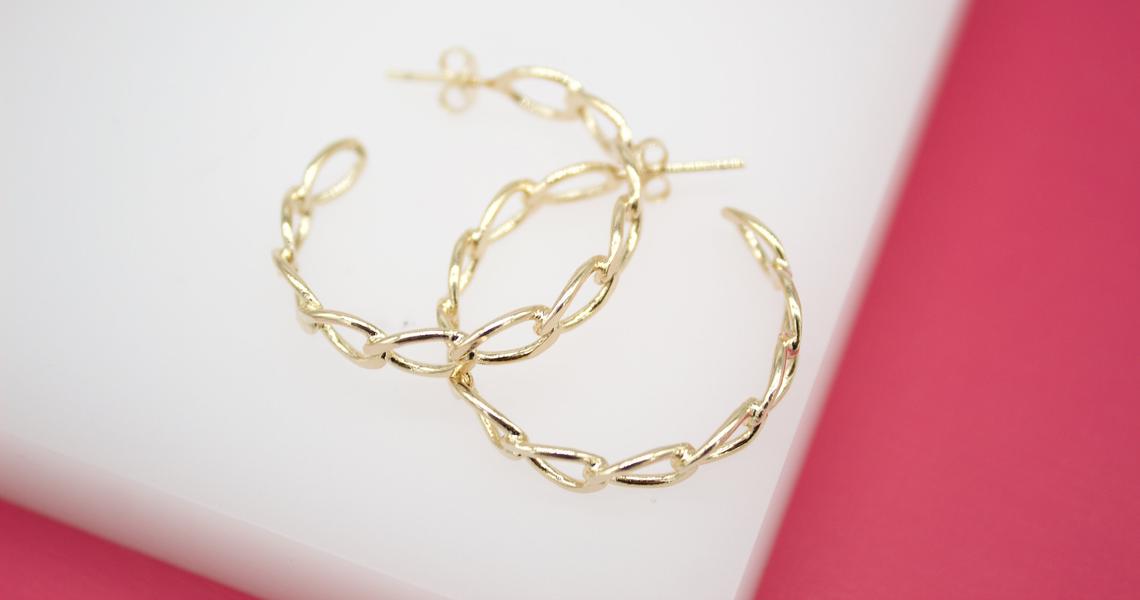 Two interlocking gold chain hoop earrings rest on a white surface with a pink background.
