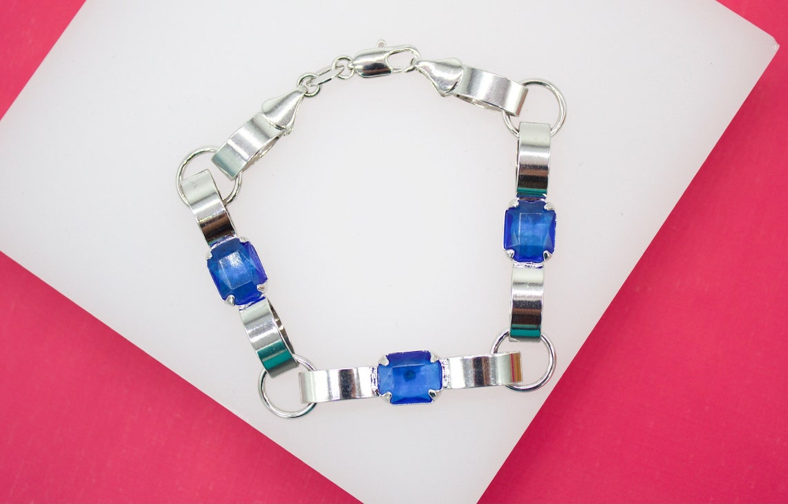 A silver link bracelet featuring vibrant blue rectangular gemstones rests on a white display surface against a pink background.
