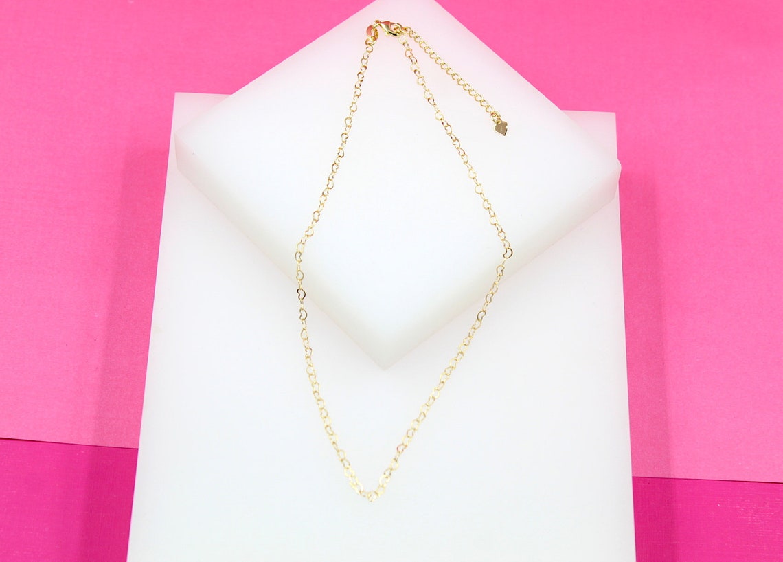 A delicate gold heart-link necklace rests on a white display block against a vibrant pink background.