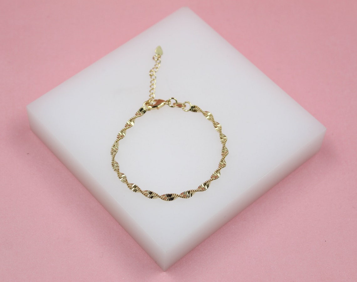 A delicate gold twisted link bracelet rests on a white display block against a pink background.