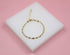 A delicate gold twisted link bracelet rests on a white display block against a pink background.