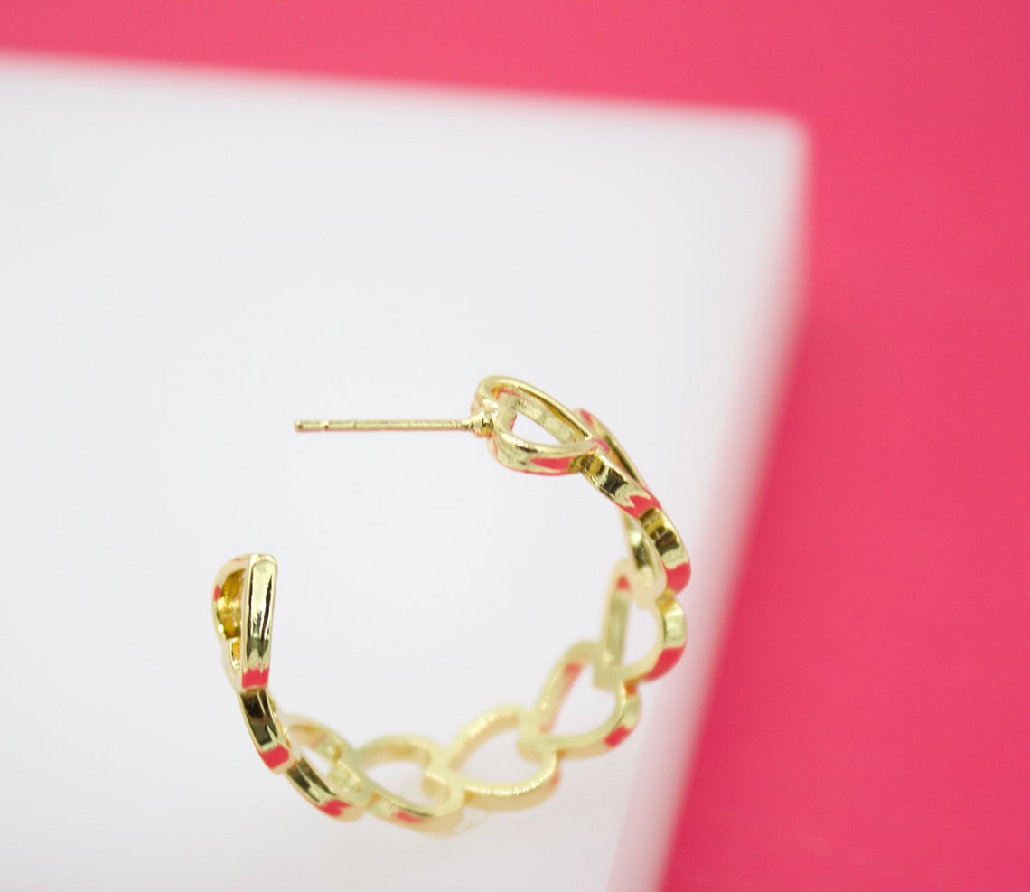A gold hoop earring featuring an interlocking heart pattern rests against a white and pink background.