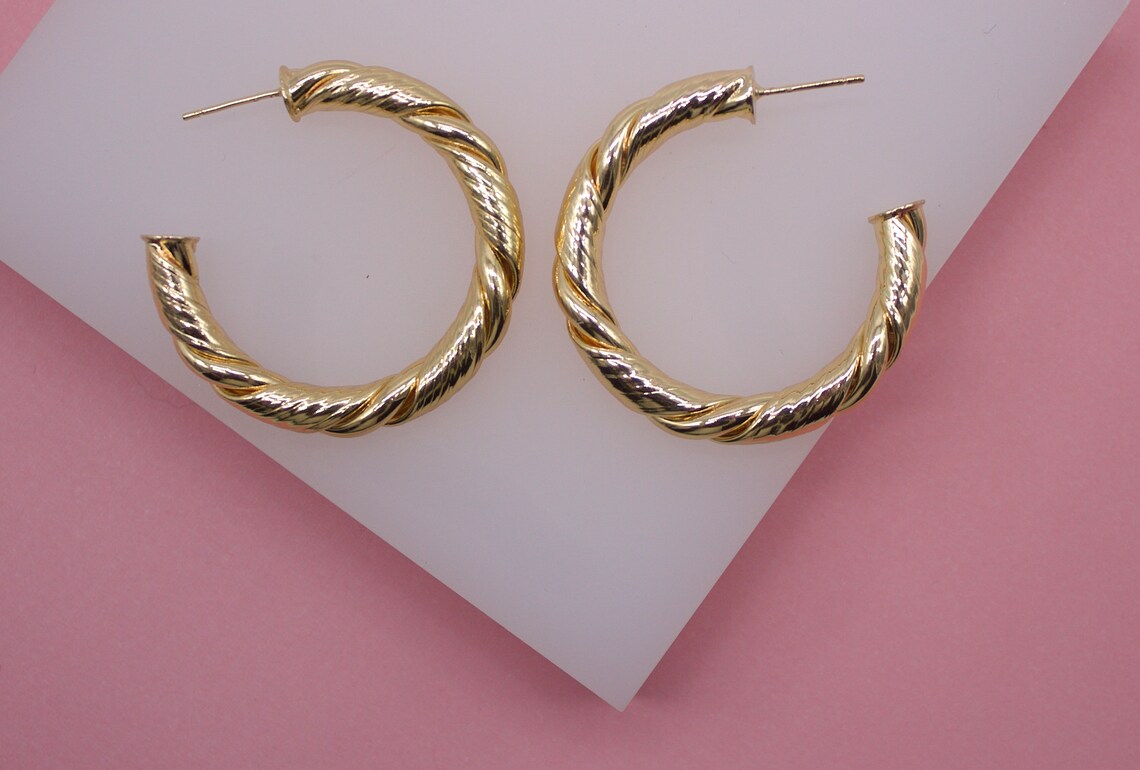 A pair of shiny gold twisted hoop earrings rest side by side on a white display stand against a pink background.