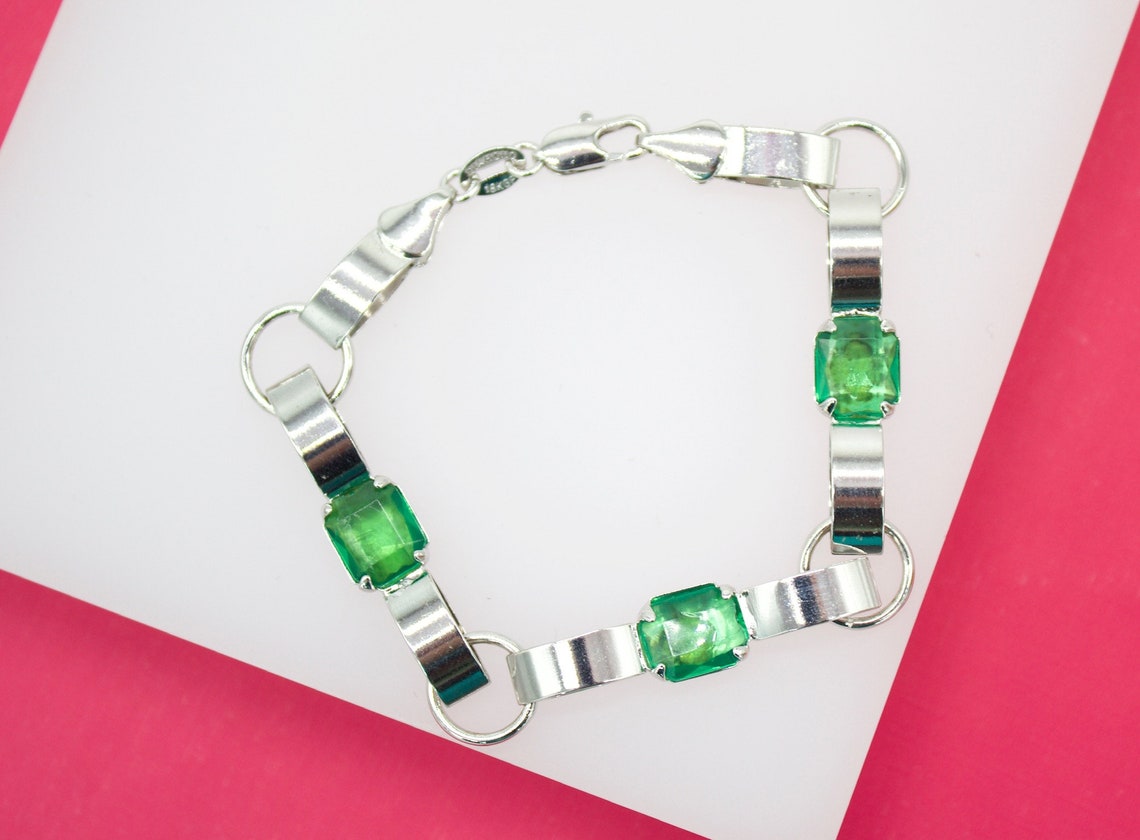 A silver link bracelet featuring vibrant emerald-green stones rests on a white and pink surface.