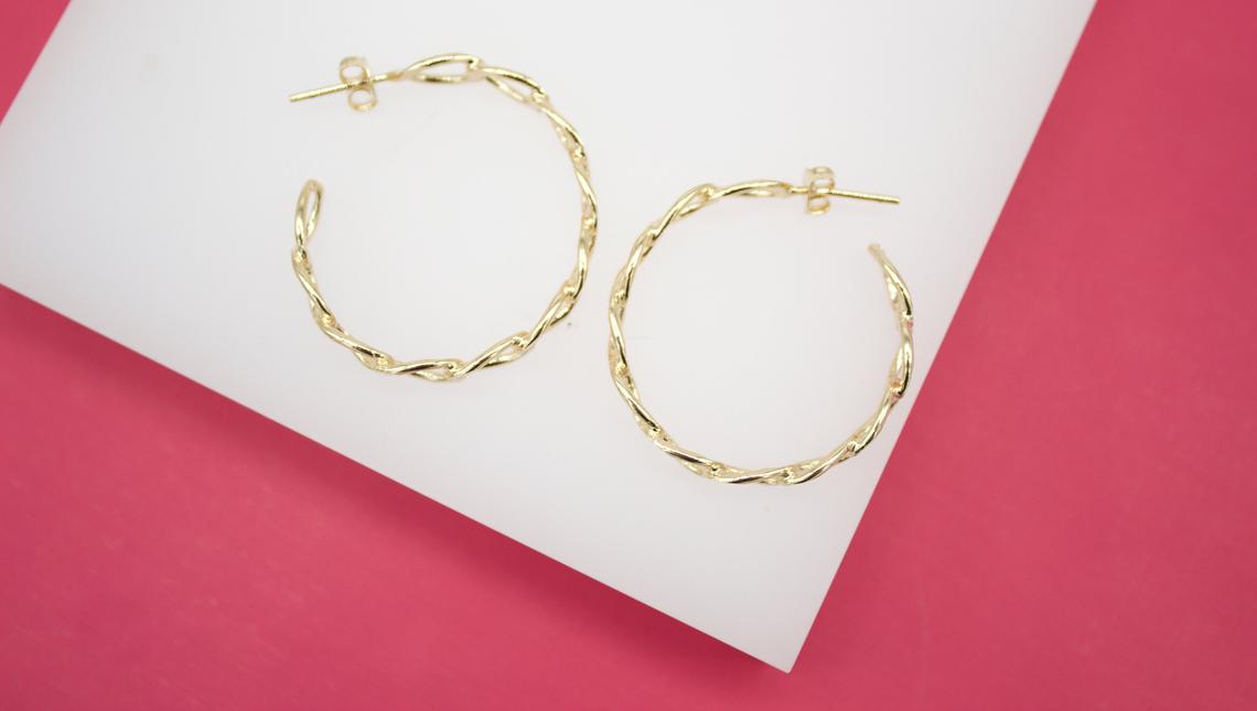 Two elegant gold twisted hoop earrings rest side by side on a white display surface against a pink background.