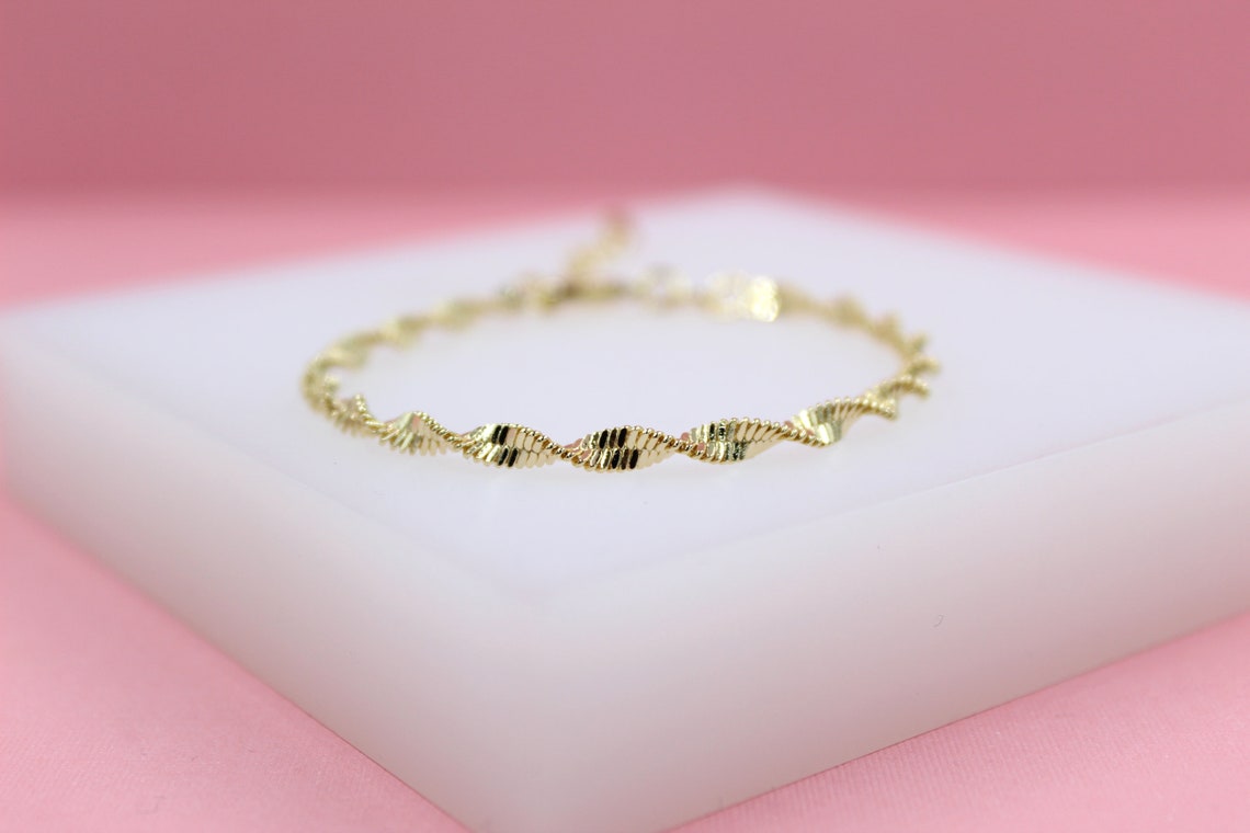 A delicate gold bracelet featuring a twisted ribbon design rests on a white display block against a soft pink backdrop.